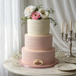 A three-tier pastel cake with textured frosting, decorated with white and pink flowers and greenery on top, displayed on a round table with candles and plates in a softly lit indoor setting.