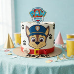 A custom white-frosted birthday cake decorated with a Paw Patrol character face, a red belt design, and a PAW Patrol topper, displayed on a decorated party table.
