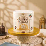 A luxury white fondant cake centered on a gold cake stand. The cake features an ornate Arabic arch design with the text "عيد مبارك في دار الأمان" in brown calligraphy, a UAE flag, a white dove in flight, and a golden Dubai skyline at the base. Colorful UAE flag brushstrokes and a gold crescent moon accent the design. The background includes warm beige tones, a decorative Arabic lantern, and soft shadow patterns. A "Gateaux" brand logo is visible on the front of the stand.