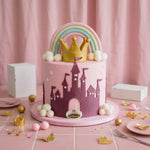 A pink and purple castle-themed birthday cake decorated with a gold crown, pastel rainbow, and white clouds, set on a pink tablecloth with party decorations.