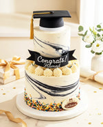 Graduation-themed cake with a marble design, 'Congrats! Hamad' banner, and decorative elements on a light background.