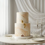 A two-tiered wedding cake with a soft blush pink and cream marble pattern and delicate gold leaf accents, displayed on a round white table with plates and a silver candelabra against a draped white fabric background.
