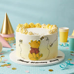 A white frosted birthday cake decorated with Winnie the Pooh illustrations and yellow whipped frosting piped around the top, displayed on a party table with paper cups, confetti, and party hats against a light blue background.
