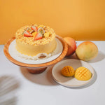 Mango cake with strawberries on a wooden stand, surrounded by mangoes on a white surface with an orange background.