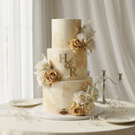 A three-tier wedding cake with a cream and gold marbled finish, decorated with white and gold roses, baby's breath, and gold monogram letters 'H&R' on the middle tier, displayed on a round table with plates and a silver candelabra against a white curtain background.