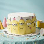 A round birthday cake decorated to look like five yellow cartoon minions with white frosting, party hats, and custom names 'Sasaad' and 'Happy Birthday Sasaad' written on the base, set on a polka-dotted tablecloth with party hats and cups.