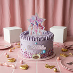 A pastel pink and lavender birthday cake decorated with purple frosting, pink sprinkles, gold foil hearts, and a star-shaped topper featuring cartoon girls and the name 'LAYAL'. The cake sits on a pink tiled table with pink plates, candy hearts, and white display blocks against a pink curtain backdrop.