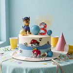 A custom 3rd birthday cake decorated with Paw Patrol characters, blue and beige balloons, and a red number 3 topper, displayed on a decorated party table.
