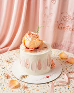 Small white cake with a pink flower on top, placed on a decorative plate with a pink curtain in the background.