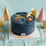 A round birthday cake decorated with a dark blue space theme, featuring small white stars, colorful planets, a seashell, and the text 'SALEM TURNING 6' and 'Happy Birthday!'. The cake sits on a white board surrounded by gold, pink, and white party cups and confetti on a light blue fabric backdrop.