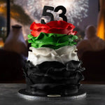Colorful cake with ruffled layers and number '53' on top, set against a blurred background of people and lights.