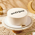 White cake with Arabic text on a decorative stand with a gold crescent moon and lantern in the background.