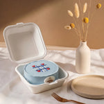 Small blue cake in a takeout container on a white surface with a vase of dried flowers in the background.