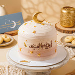 Decorative cake with gold accents on a marble stand, surrounded by cookies and a card.
