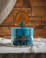 Blue cake with gold detailing and a crescent moon on top, set against a wooden wall.