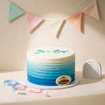 A round frosted cake with a white top and blue ombre gradient sides, decorated with the words 'It's a Boy!' in blue icing and a small oval 'Gaia's' logo badge. It sits on a white board with pastel ribbons and a white arch decoration, against a warm beige background with a soft-focus bunting banner.