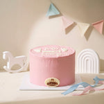 A pink frosted round cake on a white board, decorated with white icing text reading 'BABY girl landing' and a small oval logo plaque. The cake is styled on a tabletop with pastel bunting, ribbon streamers, a white rocking horse, and a white arch decoration in the background.