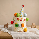 Balloon fiesta first birthday cake with "ONE" fondant letters, polka dot party hat topper, and colorful balloons and bunting