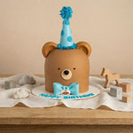 A decorated brown bear-shaped birthday cake with a blue polka-dot party hat, blue bow tie, and 'HAPPY BIRTHDAY' text on a wooden table, surrounded by small toy blocks and a wooden horse.