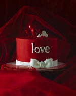 A red Valentine's Day cake with the word 'love' written on it, decorated with white ribbon and heart-shaped accents.