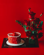 A Valentine's Day cake decorated with red and white icing, presented next to a vase of red roses, all set against a red background.