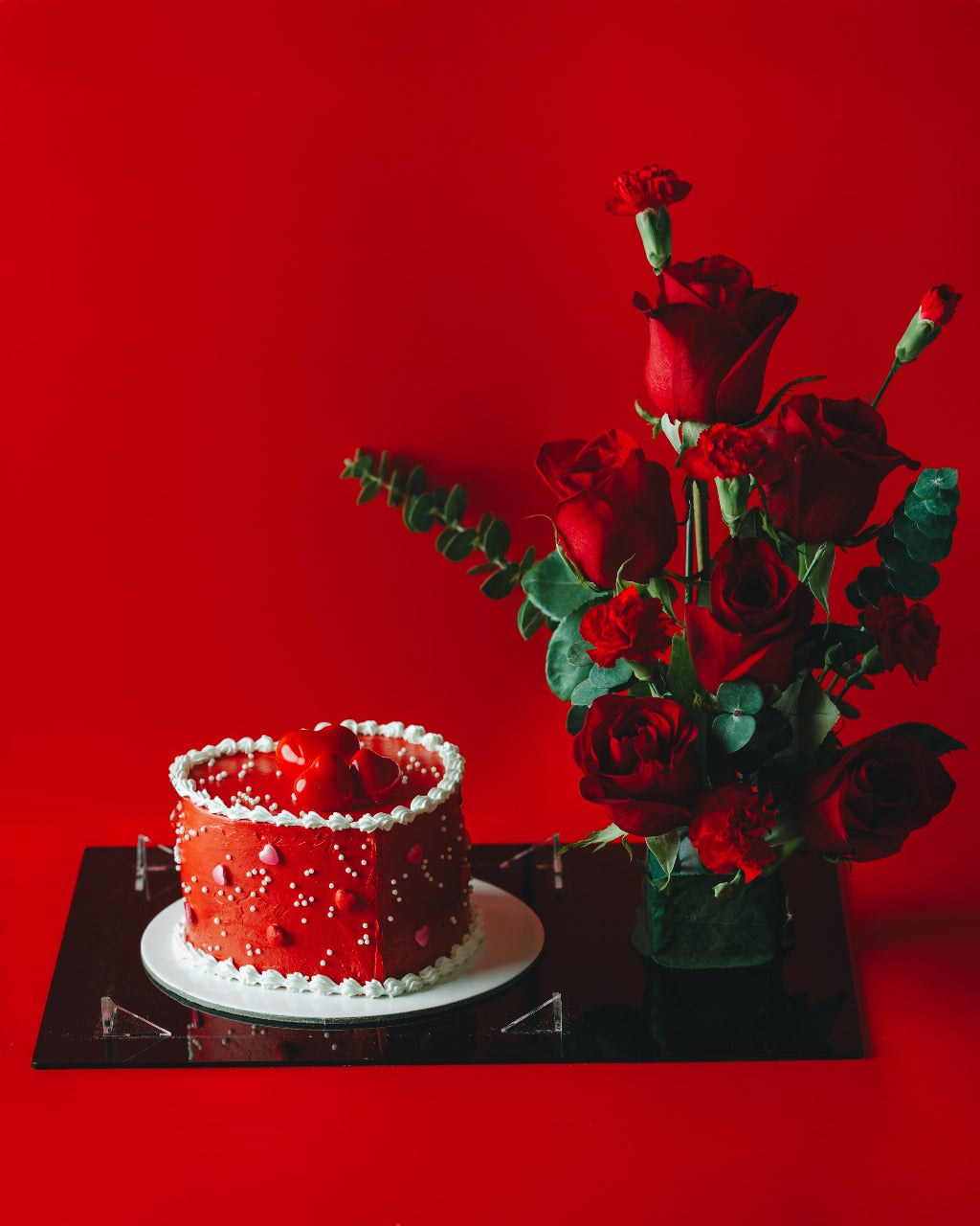 A Valentine's Day cake decorated with red and white icing, presented next to a vase of red roses, all set against a red background.