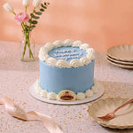 A round birthday cake with light blue frosting and white piped borders, topped with white icing script reading 'Happy Birthday anyone,' displayed on a speckled table with plates, a ribbon, flowers, and a vase in the background.