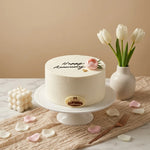 A round white frosted anniversary cake on a white pedestal stand, decorated with black script reading 'Happy Anniversary,' a small pink rose, and a gold crest plaque. The cake is styled on a marble surface with scattered pink and white petals, a cream candle, a vase of white tulips, and a fork against a warm beige background.