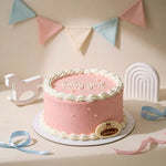 A pink baby shower cake with white piped borders and the words 'Baby girl' on top, displayed on a decorated table with pastel bunting, a rocking horse, and ribbons.