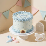 A round Baby Shower Cake with pale blue and white frosting, decorated with piped borders, small dots, and a gold oval plaque reading it's baby boy displayed on a white cake stand in a styled studio setting with pastel bunting, ribbons, and white decorative objects.