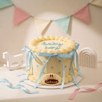A round baby shower cake with pale yellow frosting, piped cream borders, light blue ribbon bows, and blue heart accents. The top is decorated with the words 'baby boy' in light blue icing, and a round plaque at the front reads 'Gaito & Laura' with a crown logo.
