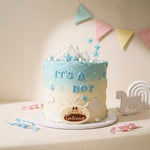A round baby shower cake with a light blue to cream-white gradient, decorated with clouds, stars, and the words 'IT'S A BOY' in icing. The cake sits on a white board with a round logo plaque that reads 'Gateaux'.