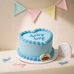 A light blue heart-shaped cake with piped swirl borders and the words 'happy birthday' written on top, displayed on a white round board with a small cake topper. The scene is styled with pastel bunting, bows, and a small rainbow decoration against a warm cream background.