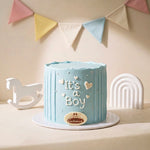 A light blue frosted Baby Boy Shower Cake 'It's a Boy' and a small crown decoration, displayed on a white round cake board with a white rolyoly and a white arch decoration in the background, under a pastel bunting banner.