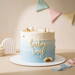 A round frosted cake with a light blue to white ombre design, decorated with gold and white beads, gold lettering that reads 'baby boy', and a small oval cake topper logo. The cake sits on a white round board on a beige table with pastel bunting in the background.