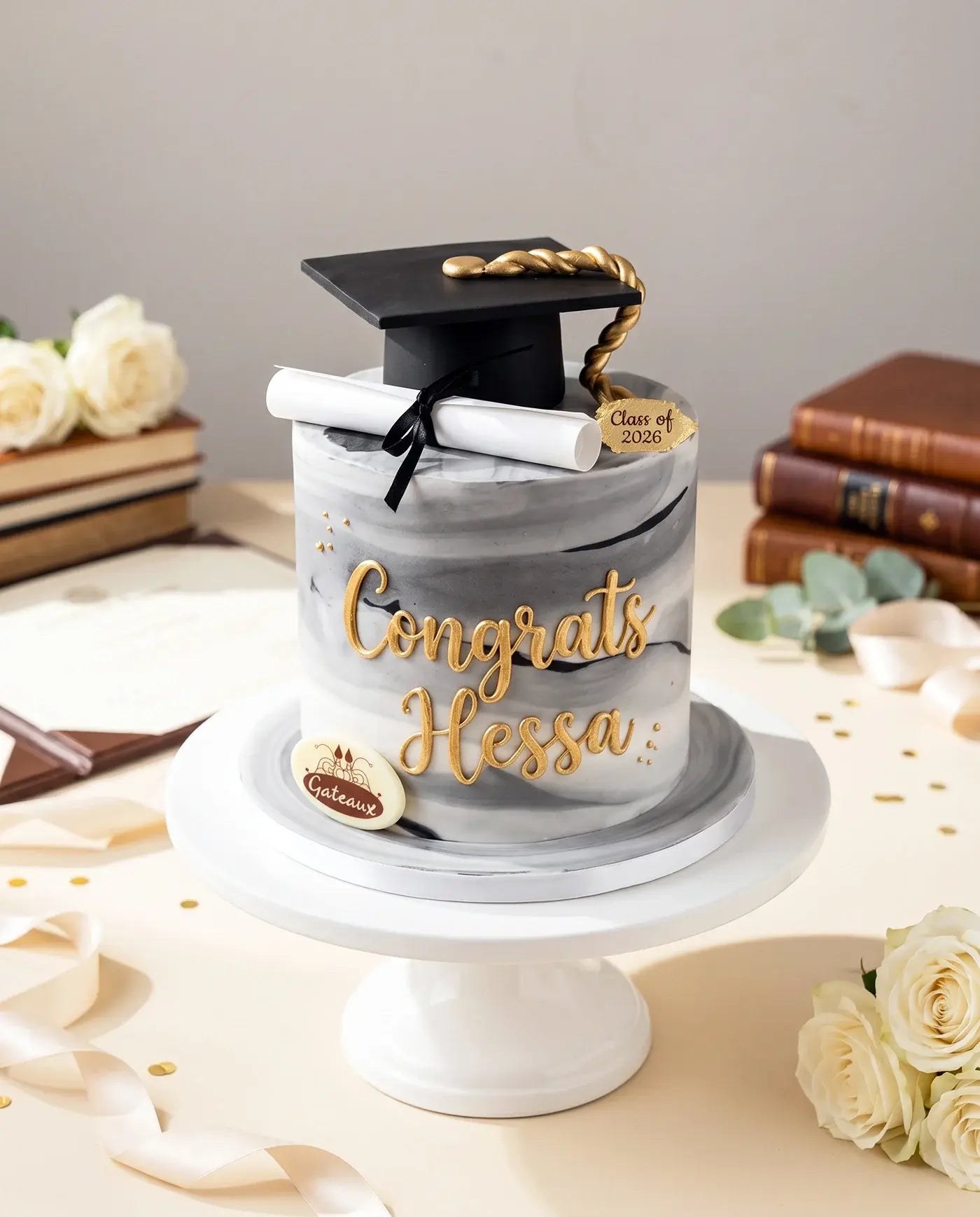 Graduation-themed cake with a mortarboard and diploma on a white stand, surrounded by flowers and books.