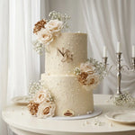 A two-tier cream wedding cake decorated with white roses, gold rose accents, baby's breath, and a gold 'N & S' monogram, displayed on a white table with a candelabra in the background.