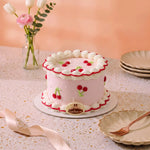 A pink frosted cake decorated with white whipped borders, red cherries, and green leaf accents, displayed on a decorated table with plates, a ribbon, and flowers in the background.