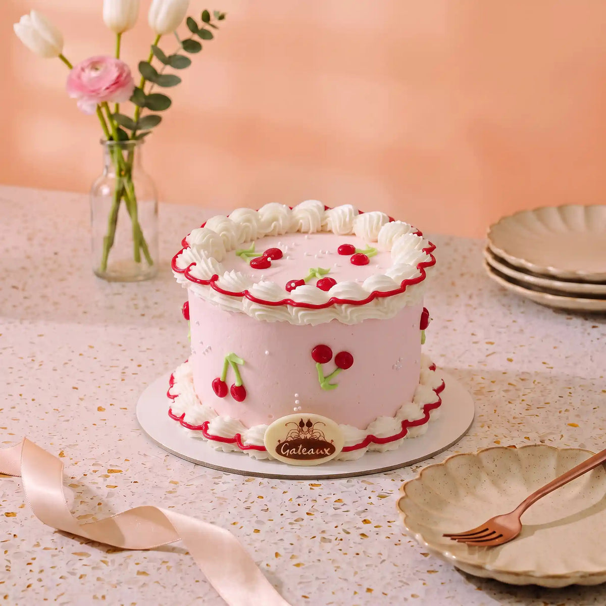 A pink frosted cake decorated with white whipped borders, red cherries, and green leaf accents, displayed on a decorated table with plates, a ribbon, and flowers in the background.