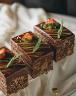 A chocolate cake slice topped with a strawberry and a sprig of mint, garnished with decorative chocolate shavings.