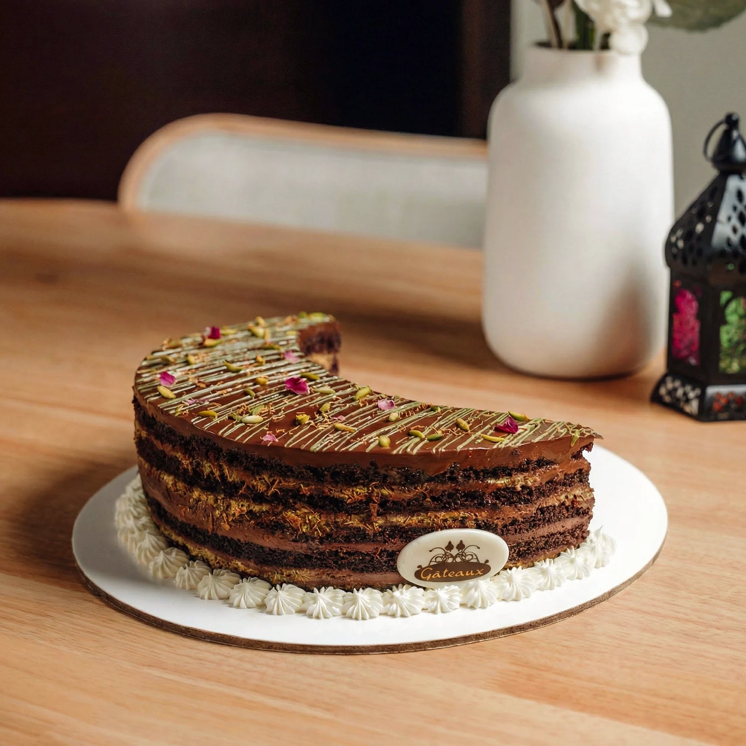 Chocolate cake with white frosting on a wooden table with a vase and lantern in the background.