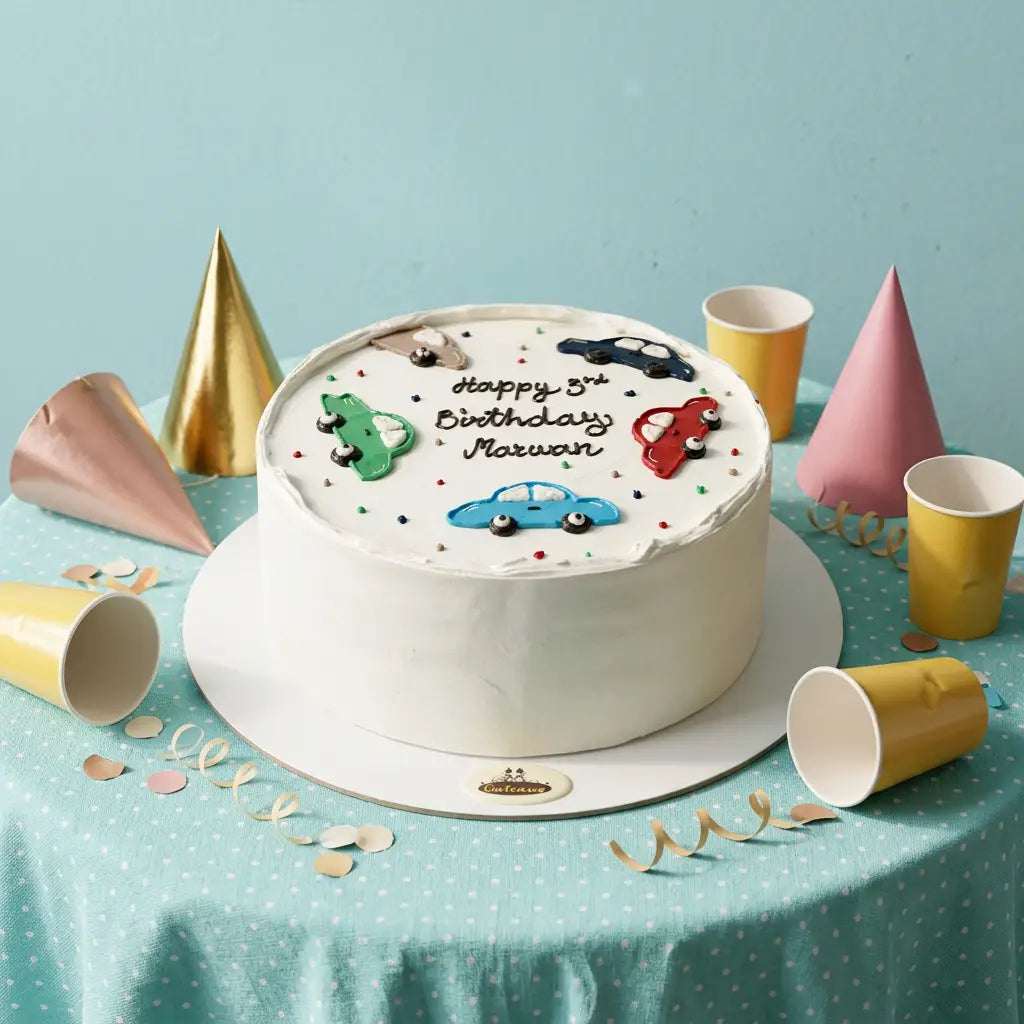 A round white-frosted birthday cake with car decorations and the text 'Happy 5th Birthday Maxwan' sits on a table covered with a light blue polka-dot cloth. Gold, pink, and yellow party hats, cups, and confetti are arranged around it against a light blue background.