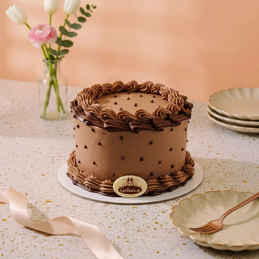 A round chocolate cake with piped frosting borders and chocolate dots, displayed on a speckled table with plates, a ribbon, and flowers in the background.