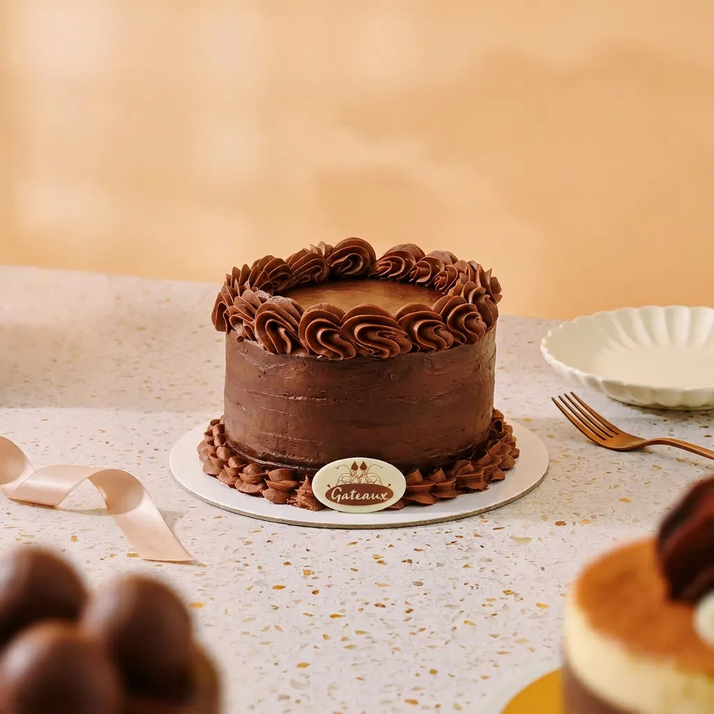 A chocolate cake with piped frosting rosettes on top and around the base, displayed on a white plate on a speckled countertop. A small round plaque in front reads Gauthier, and a fork and plate are visible nearby.
