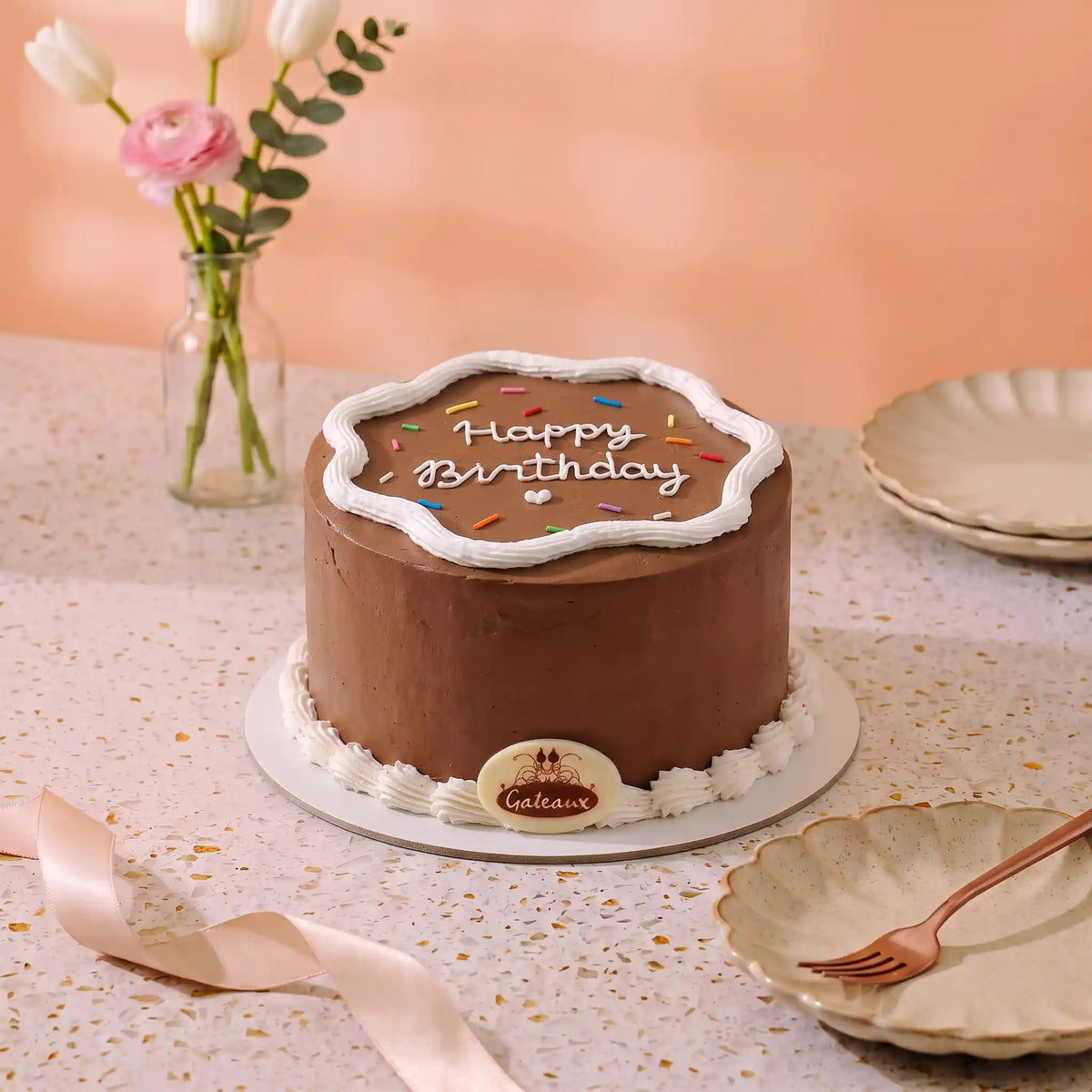 A round chocolate cake with white frosting, colorful sprinkles, and a white piped border on top, decorated with a small gold cake topper reading 'Goldluxe' and 'Happy Birthday.' The cake sits on a speckled table with plates, a ribbon, flowers, and a peach-toned background.