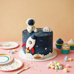 A round cake decorated with a space theme, featuring a dark blue top, gold stars, white clouds, a white astronaut topper, and a rocket decoration. The cake sits on a pink table surrounded by pastel cupcakes, plates, and candy against a peach background.