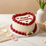 A heart-shaped anniversary cake with white frosting, red piped borders, and 'Happy Anniversary' written in red icing on top, displayed on a marble surface with decorative petals, a vase, and grapes in the background.