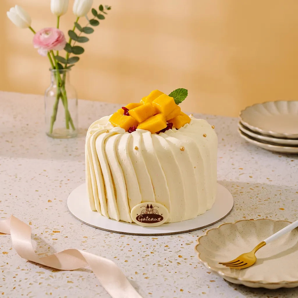 A frosted round cake on a white board, decorated with yellow mango pieces and a mint leaf, set on a speckled tabletop with flowers and plates in the background.