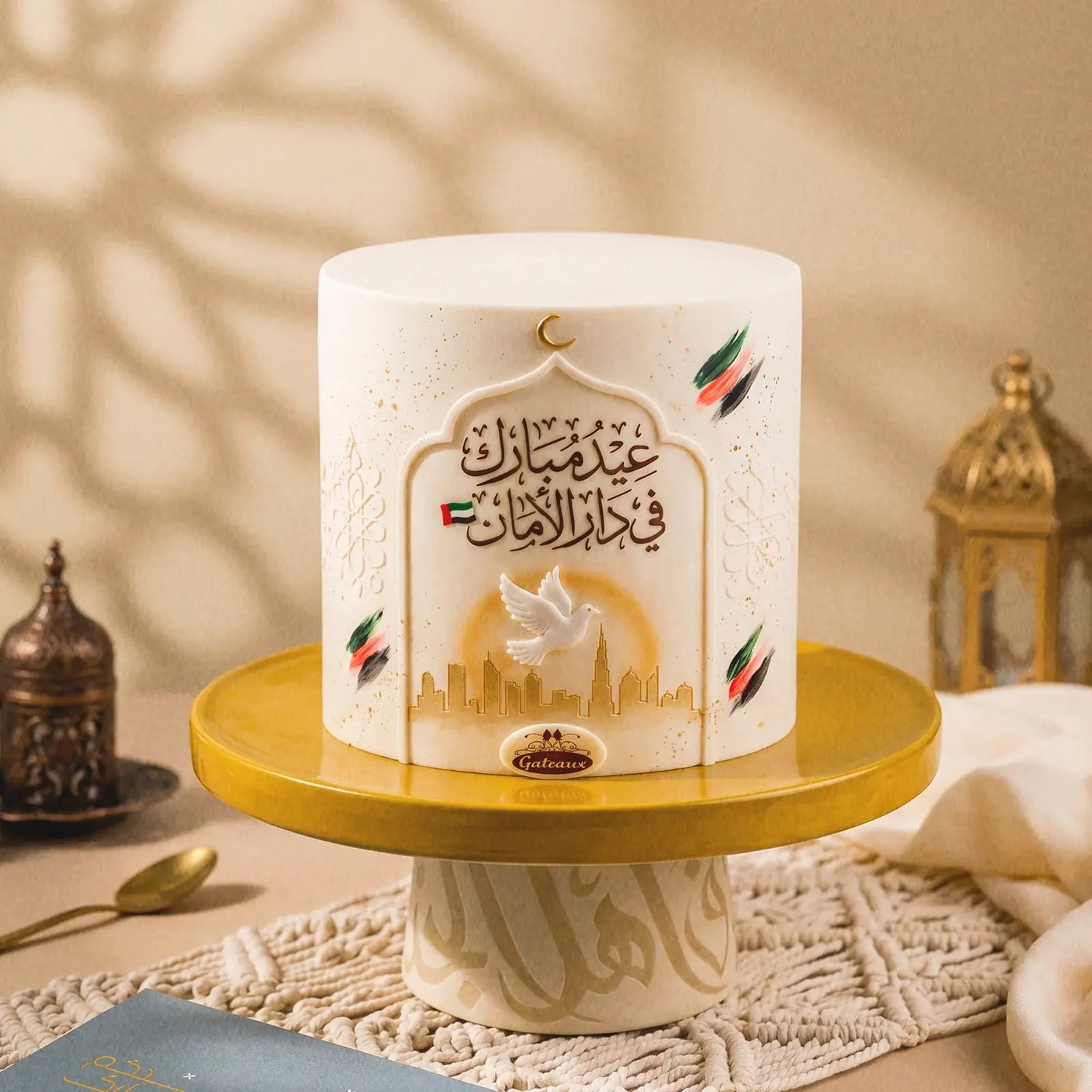 A luxury white fondant cake centered on a gold cake stand. The cake features an ornate Arabic arch design with the text "عيد مبارك في دار الأمان" in brown calligraphy, a UAE flag, a white dove in flight, and a golden Dubai skyline at the base. Colorful UAE flag brushstrokes and a gold crescent moon accent the design. The background includes warm beige tones, a decorative Arabic lantern, and soft shadow patterns. A "Gateaux" brand logo is visible on the front of the stand.

