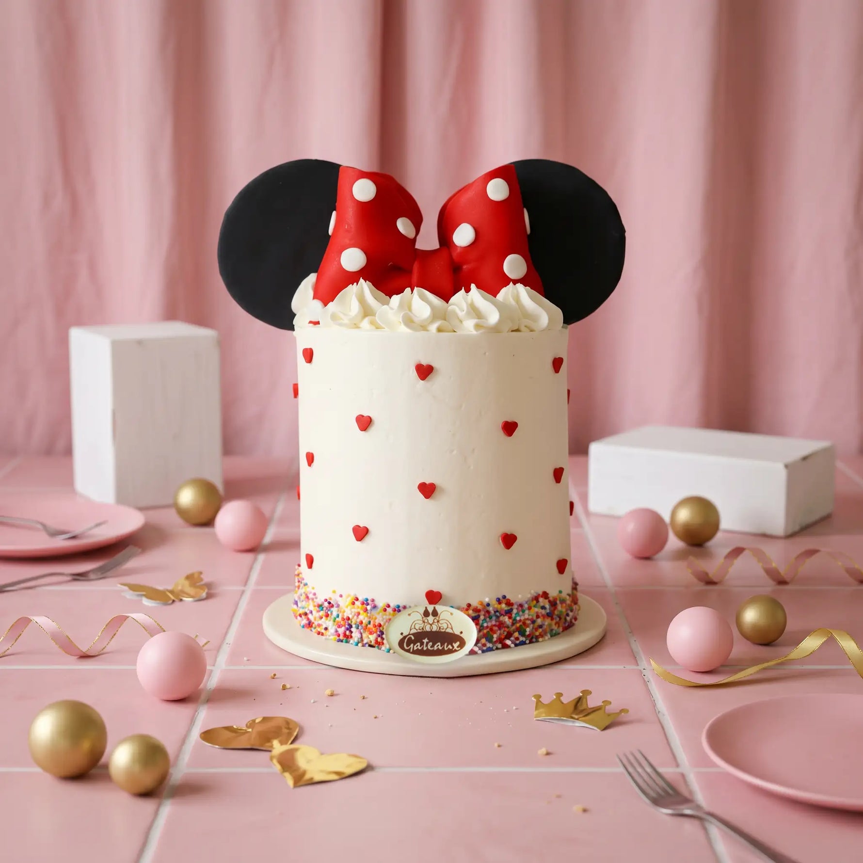 A Minnie Mouse-themed birthday cake with white frosting, red and white polka-dot bow topper, black mouse ears, and small red heart decorations, set on a pink tablecloth with pink plates, gold and pink eggs, and ribbon props.